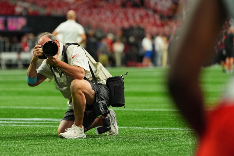 A photographer kneels on a football field, aiming a large camera lens. He's wearing a light shirt, shorts, and has a camera bag. The background shows blurred spectators in a stadium.