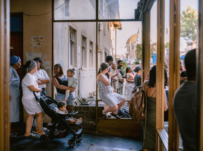A diverse group of people, including women, children, and babies in strollers, gather and interact outside a building on a sunny day, some standing and some sitting on ledges or steps.