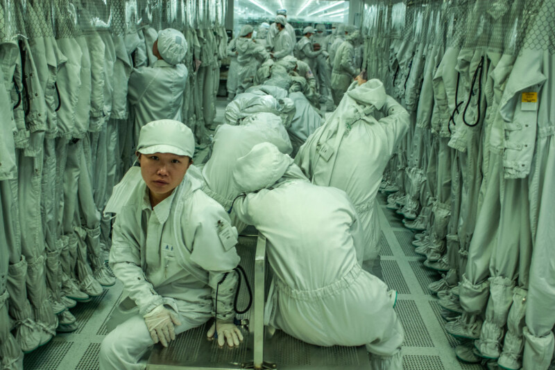 Workers dressed in white protective suits and caps sit and crouch in a cleanroom surrounded by rows of hanging suits, with one person looking directly at the camera in a brightly lit, sterile environment.