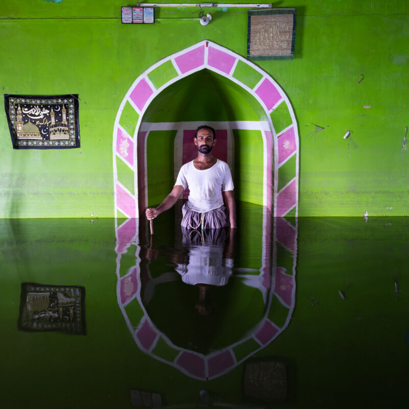 A man stands waist-deep in floodwater inside a vividly painted green room with a pink-arched doorway and Islamic wall hangings, his reflection visible in the water.