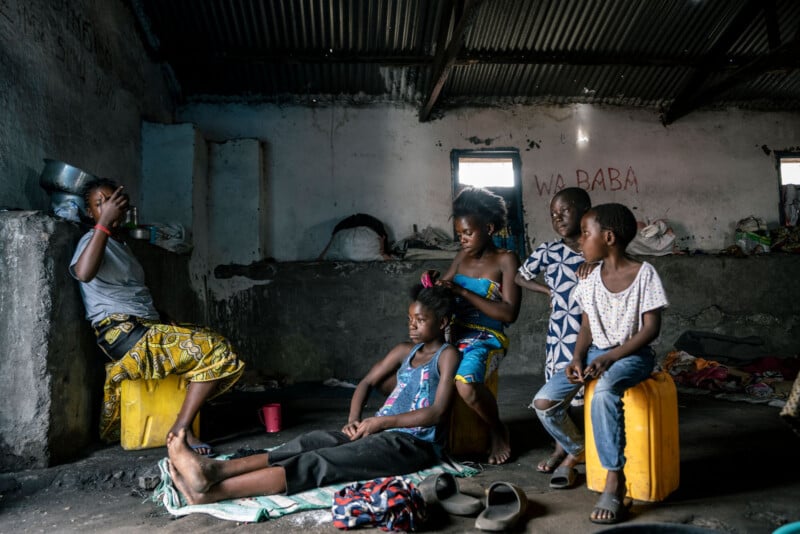 Five children sit and relax inside a dimly lit, worn room with concrete walls. One child drinks from a metal bowl, while others sit or lounge on yellow containers and fabrics. Sunlight enters through small windows.