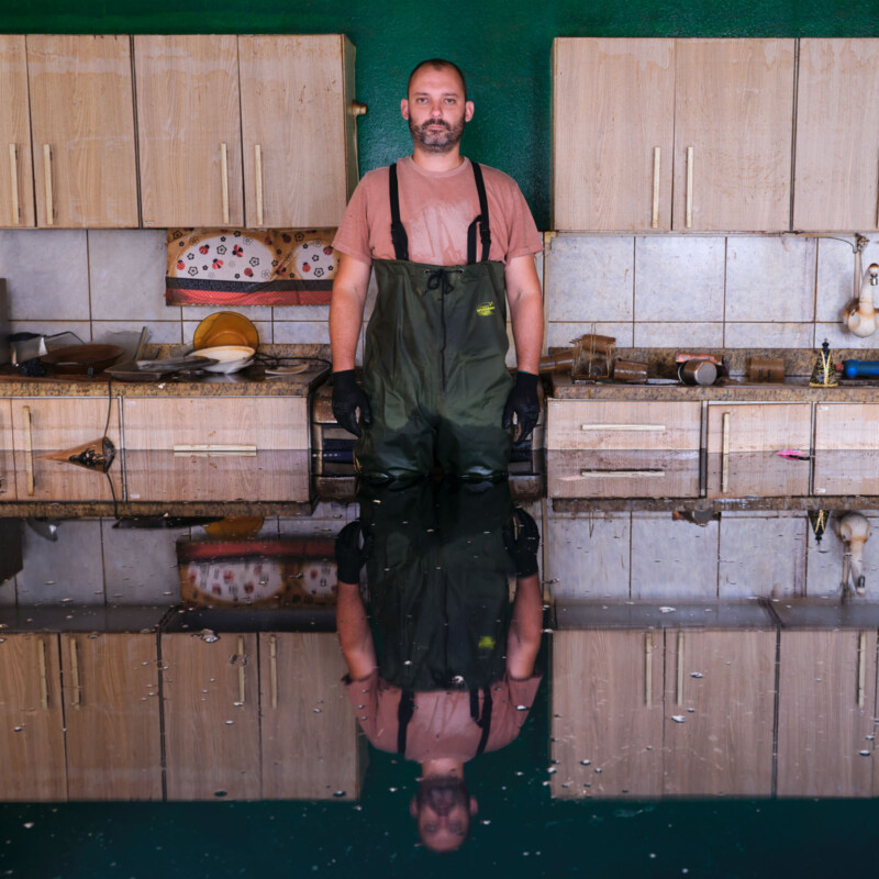 A man in waterproof overalls stands in a flooded kitchen with dirty dishes on the counter; water covers the floor, creating his clear reflection.
