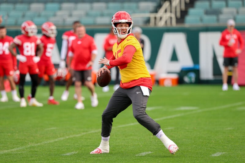 A football player in a yellow jersey and gray pants prepares to throw the ball during practice on a grassy field. Other players in red uniforms and coaches are in the background. The setting appears to be a stadium.