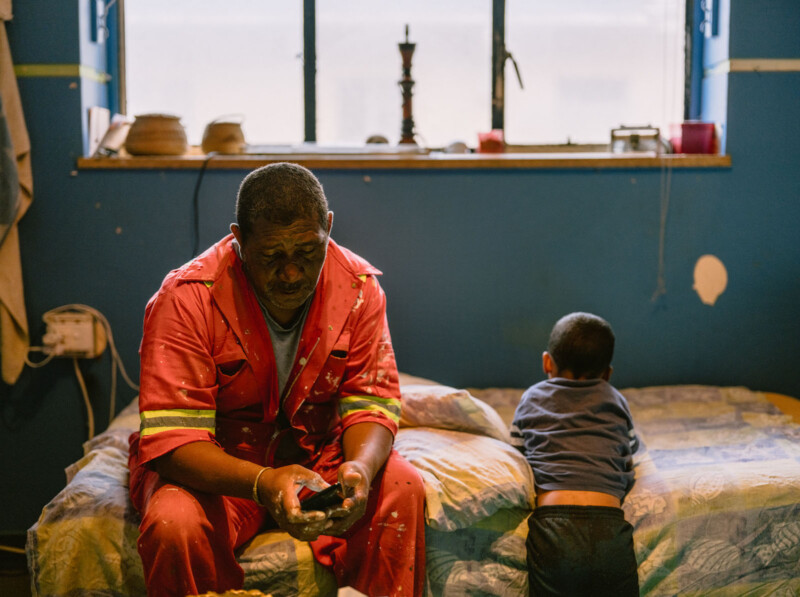 A man in a red jumpsuit sits on a bed looking at his phone, while a young boy leans over the bed facing away. The room has a blue wall, a window with objects on the sill, and soft natural light.