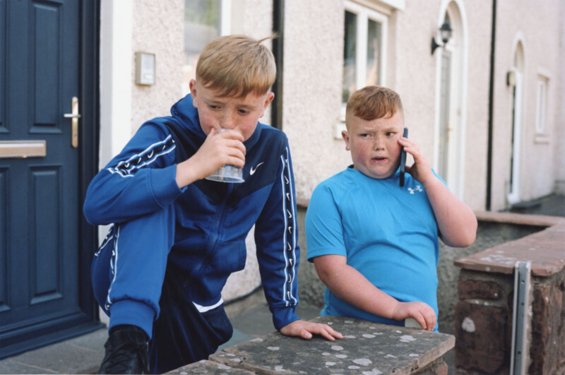 Two boys stand outside a house. One boy in a blue tracksuit drinks from a glass with one leg on a low wall, while the other boy in a light blue shirt talks on a phone, looking slightly to the side.