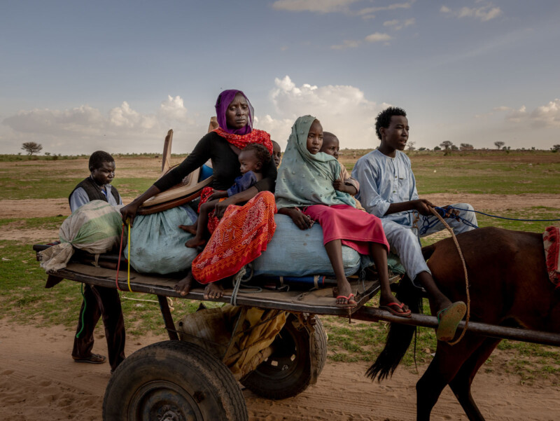 A family rides on a wooden cart pulled by a horse through a rural landscape, with women and children sitting on bundles and a man guiding the cart.