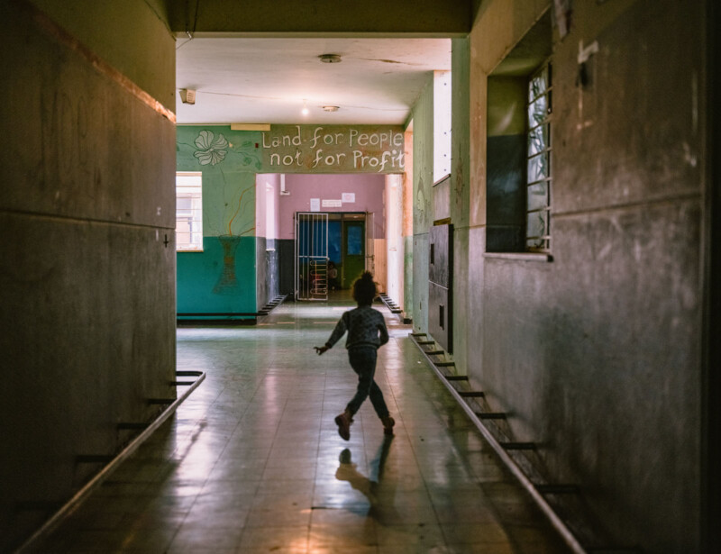 A child runs down a dimly lit hallway toward a colorful wall with the words "Land for People not for Profit" painted on it. Light from windows illuminates the corridor.