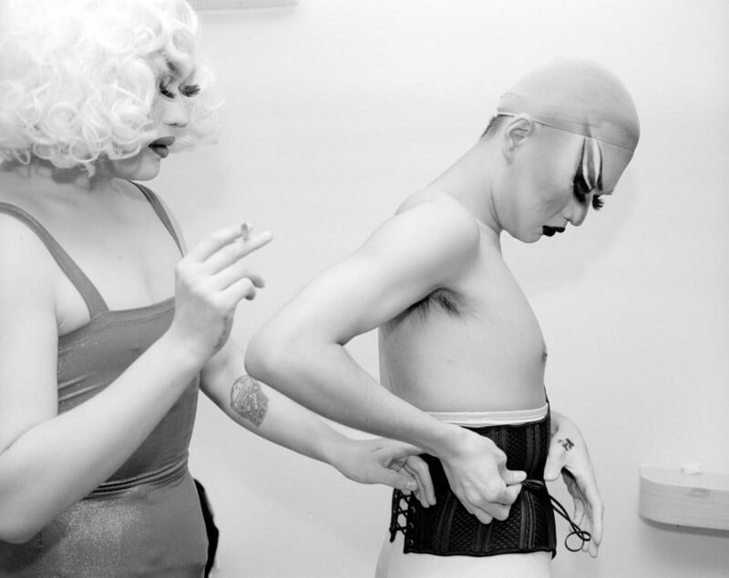 Two people in a dressing room; one in a wig and dress holds a cigarette while helping the other, who is shirtless and wearing a bald cap and dramatic makeup, tighten a corset with laces.