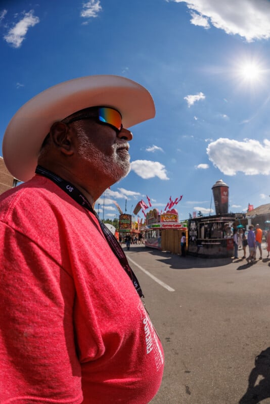 A man in a red shirt, white cowboy hat, and reflective sunglasses stands outdoors at a fair, with food stands, flags, and people in the background under a sunny, blue sky with scattered clouds.
