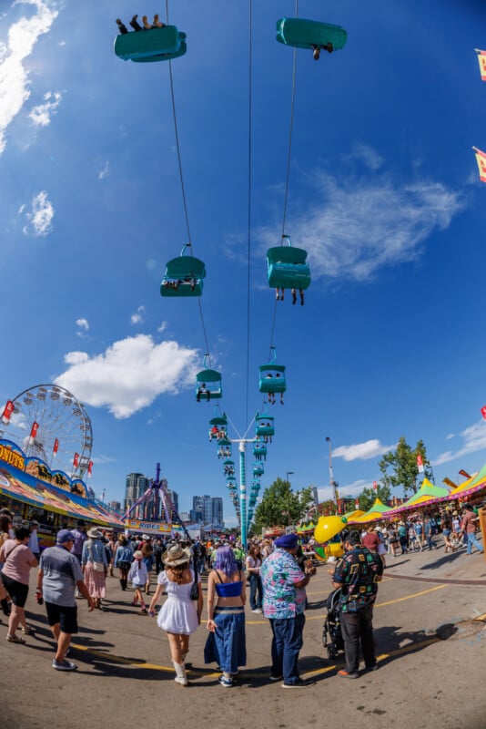 People walk through a busy fairground, with a ferris wheel and colorful stalls on each side. Above, teal cable cars with passengers ride across a bright blue sky dotted with clouds.