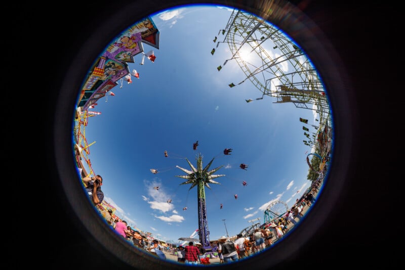 A fisheye lens captures a vibrant carnival scene with a tall swing ride at the center, people flying in chairs, blue sky above, and colorful booths and crowds circling the frame’s edge.