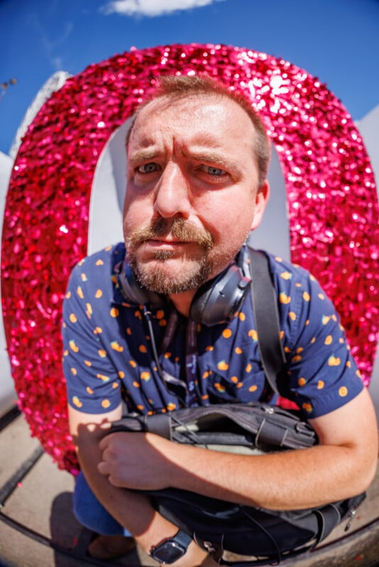 A man with a beard and mustache, wearing a blue shirt with orange dots and headphones around his neck, crouches in front of a large pink glittery letter "O" outdoors under a blue sky.