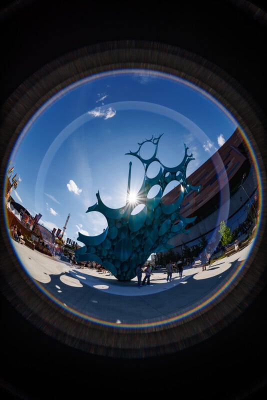 A fisheye lens photo of a modern outdoor sculpture made of blue circular shapes, with sunlight shining through and people walking nearby. Surrounding buildings and a blue sky with clouds are visible. Rainbow lens flares encircle the image.