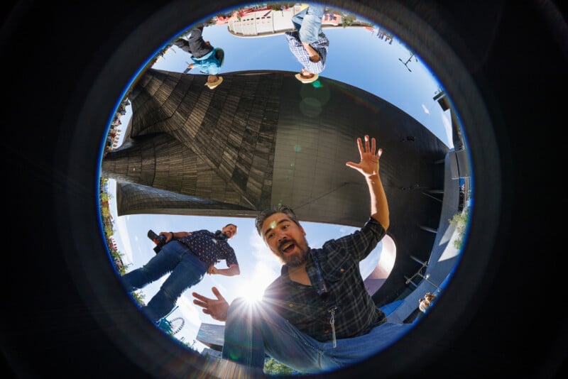 A man with a surprised expression leans toward a fisheye lens, hands raised, as another person stands behind him. The scene is outdoors near a large modern sculpture, with sunlight creating a starburst effect.