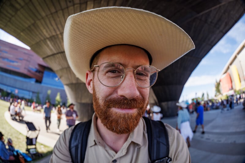 A bearded man wearing glasses and a wide-brimmed hat smiles at the camera. He is outdoors at a public event, with people and buildings in the background, photographed with a fisheye lens.