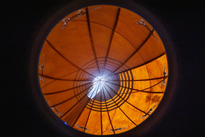 View from inside a circular structure with an orange canopy and geometric patterns, featuring silhouettes of lizards. Light shines through the center, creating a bright focal point and radiating lines.