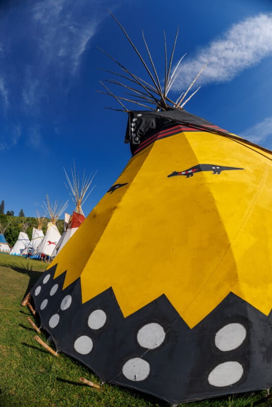 A bright yellow tipi with black and white painted designs stands on grass under a blue sky, with other colorful tipis visible in the background.