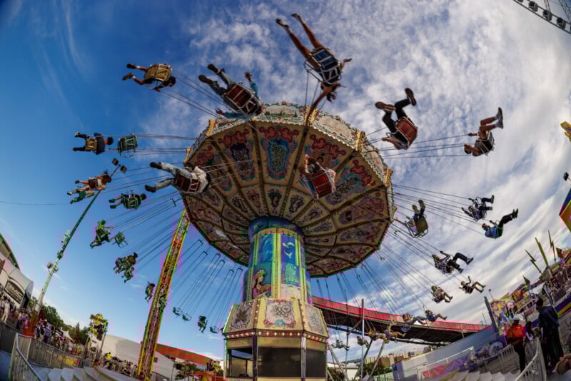 People ride a colorful swing carousel at an amusement park, with seats flying outward as the ride spins. The sky is clear and blue, adding to the lively, festive atmosphere.