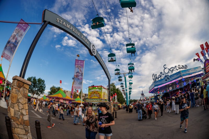 A lively scene at a fairground with crowds walking, a large "Stampede Park" arch, colorful rides, food stands, and people riding a blue sky tram under a partly cloudy sky.