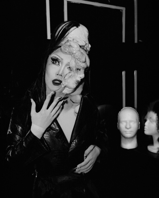 A person wearing dramatic makeup and a black leather outfit poses with a surreal mask-like sculpture partly covering their face. Mannequin heads with wigs are visible in the dimly lit background. The image is in black and white.