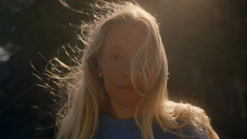 A woman with long blonde hair stands outdoors in soft sunlight, wearing a blue sweater. Wind blows her hair across her face, and the background is softly blurred with natural, dark green tones.