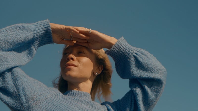 A woman wearing a blue sweater shields her eyes from the sun with both hands, looking into the distance against a clear blue sky.