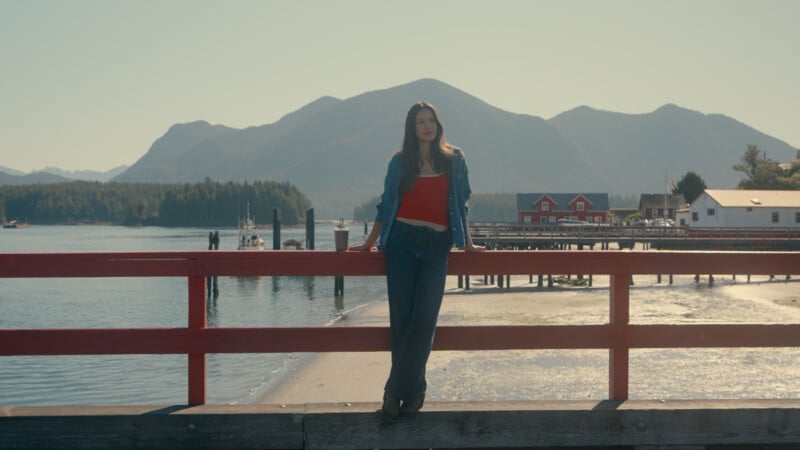 A woman in a red top and jeans stands on a pier with red railings, with mountains, water, and buildings in the background under a clear sky.