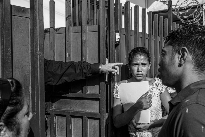 A young woman clutching papers stands in front of a gate, looking concerned. A man’s hand points at her from behind the gate, while two other people stand nearby in a tense scene. The photo is in black and white.
