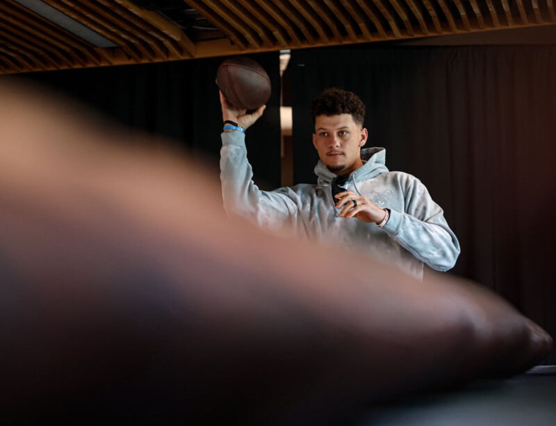 A person wearing a light gray hoodie holds a football in a throwing position, focused on the unseen target. The foreground is softly blurred, with wooden ceiling panels and a dark curtain in the background.