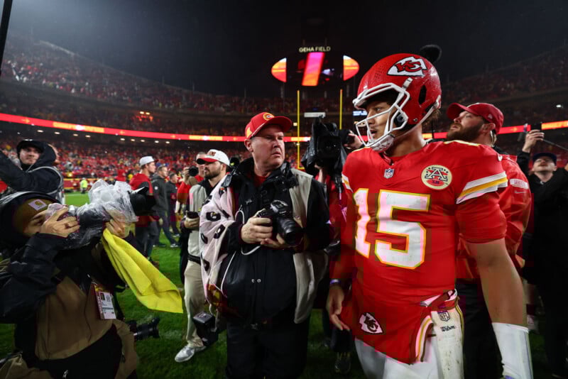 A football player in a red jersey with the number 15 celebrates on the field. He is surrounded by photographers and media personnel. The stadium is filled with fans, and a large screen is visible in the background.