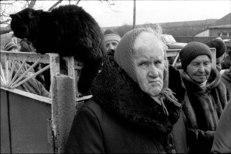 An elderly woman in a headscarf stands outdoors among a group of people, looking serious. A black cat sits on a fence nearby. The scene appears to be in a rural area on a cold day.