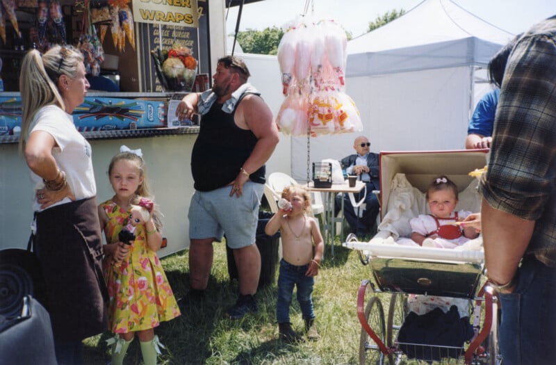 A group of people at an outdoor fair: two young girls, one holding a doll, a shirtless small boy, a baby in a stroller, and several adults near a food stall and tents, with cotton candy hanging above.