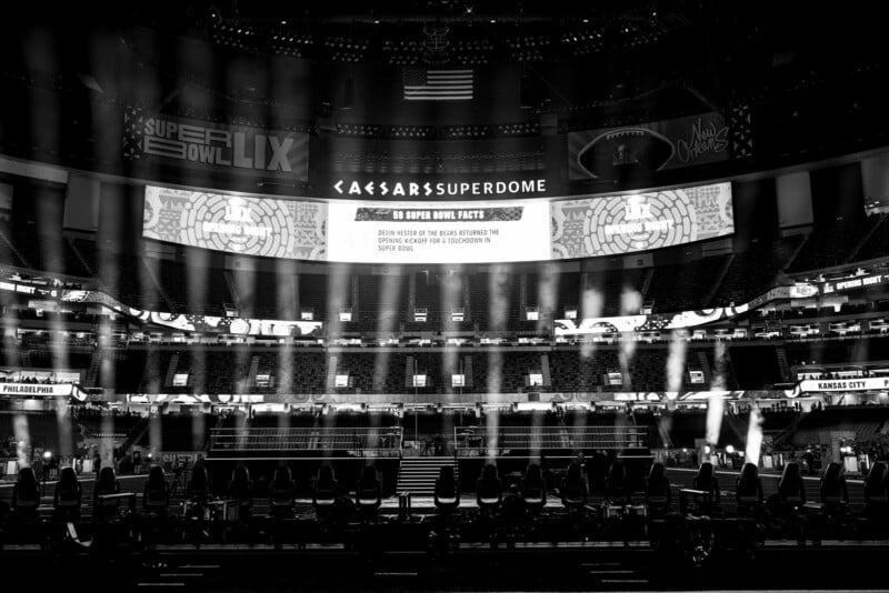 Black and white image of an empty sports stadium, with "Caesars Superdome" on the scoreboard. Spotlights illuminate the field, and there are banners displaying "Super Bowl LIX." A large screen shares football-related facts.