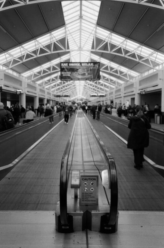 A black-and-white photo of a busy airport terminal shows people walking on either side of a moving walkway under a high, geometric skylight ceiling. A caution sign is visible at the start of the walkway.