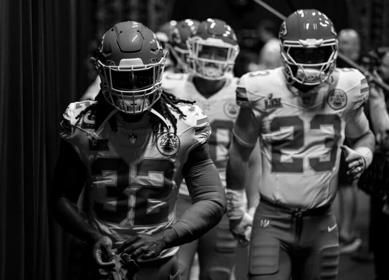A group of American football players in uniform walking through a tunnel, led by a player wearing jersey number 32. They are wearing helmets and football gear. The image is in black and white.
