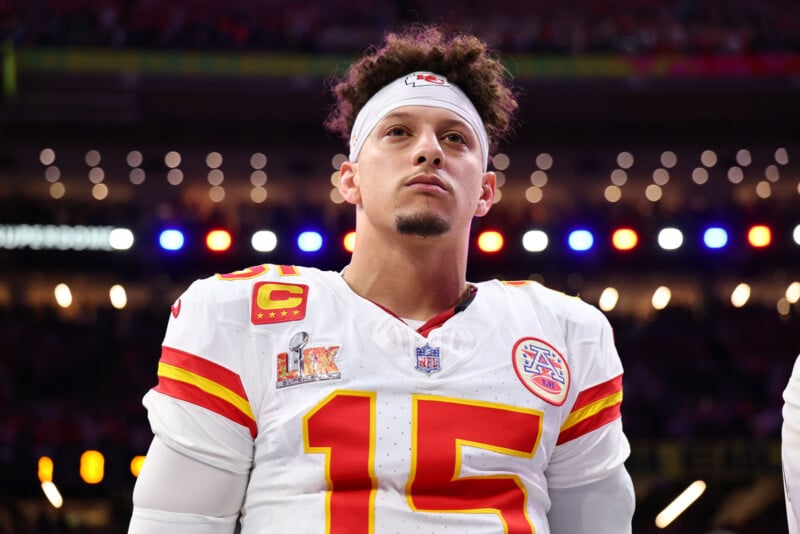 A football player in a white and red jersey with the number 15 stands focused under stadium lights. He wears a white headband and has curly hair. The background shows blurry lights and a partial view of the audience.