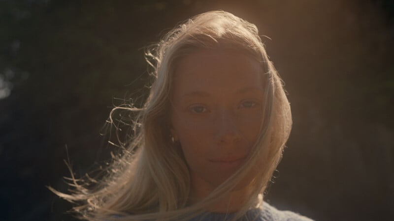 A woman with long blonde hair stands outdoors, illuminated by warm sunlight. Some hair gently blows across her face as she gazes calmly at the camera, with a dark, blurred background behind her.