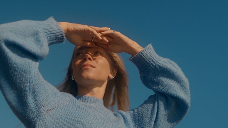 A woman in a light blue sweater stands outdoors, shielding her eyes with her hand as she looks up towards the sky on a bright, sunny day. The clear blue sky serves as the background.