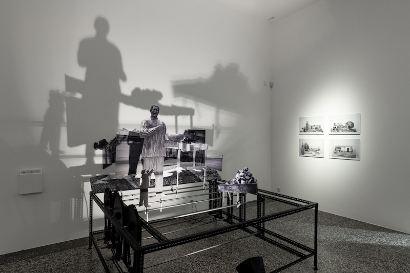 A contemporary art installation in a gallery features monochrome cutouts of a person and objects on a black metal frame, casting large shadows on a white wall. Black-and-white photographs are displayed on the adjacent wall.
