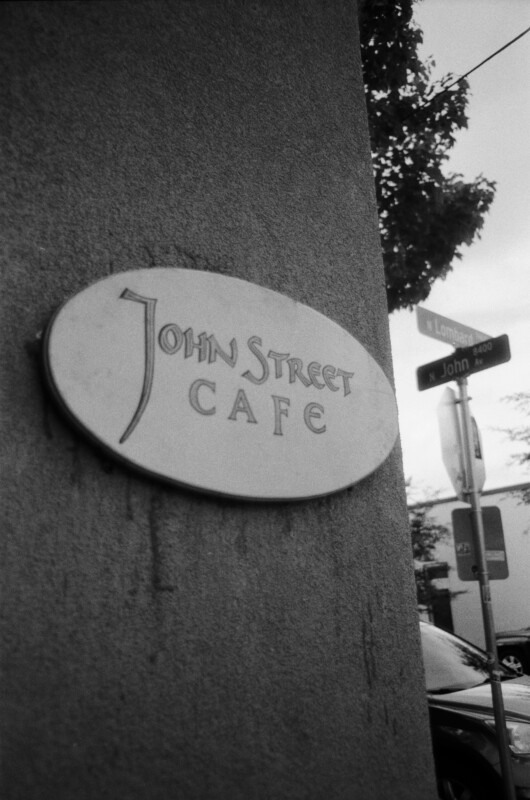 A sign reading "John Street Cafe" is mounted on a wall. In the background, there are street signs for N Lombard St and N John Ave, parked cars, and some trees. The photo is in black and white.