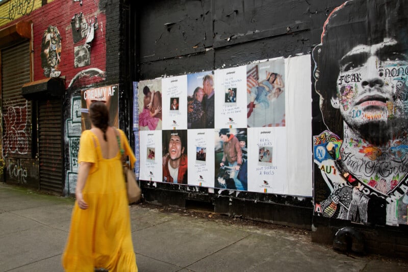 A woman in a yellow dress walks past a graffiti-covered wall with posters of people in various poses, including close-ups and candid shots. The wall also features a large, detailed mural of a man’s face.
