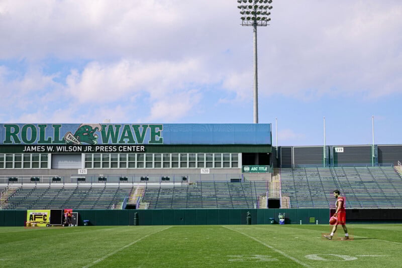 A person in a red outfit walks across a football field. The stands are empty, and a large sign reads "ROLL WAVE" at the James M. Wilson Jr. Press Center. The sky is partly cloudy.
