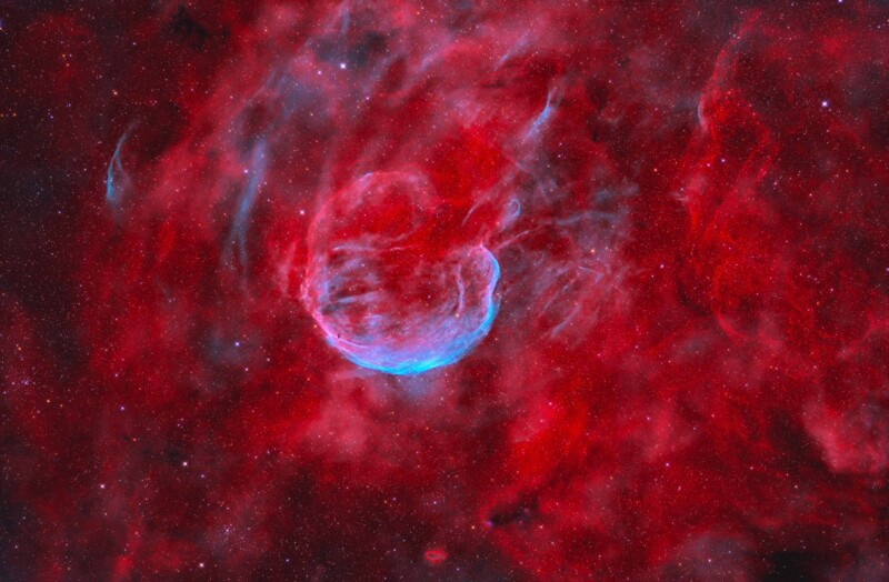A vibrant nebula in space, featuring a blue, bubble-like structure surrounded by swirling red gas clouds and scattered stars on a dark background.