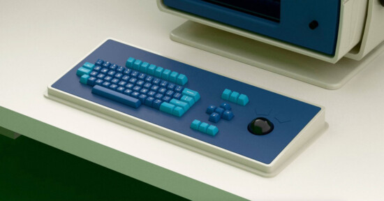 A retro-style computer keyboard with blue and turquoise keys, an unusual layout, and a built-in black circular trackpad, on a white desk next to a matching computer monitor.