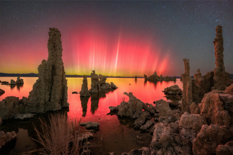 Tall, jagged tufa rock formations rise from the calm waters of Mono Lake at night, illuminated by vibrant red and pink aurora lights in the sky, with stars visible above.