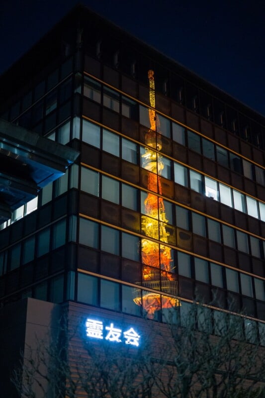 A building with a reflective glass facade shows the illuminated image of Tokyo Tower at night. The glow of the tower creates a vibrant pattern on the building. A neon sign with Japanese characters is visible below.