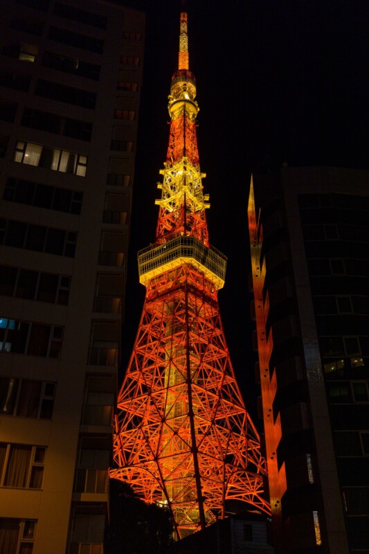 Tokyo Tower illuminated at night, glowing in bright red and orange lights. The metallic structure is framed by two dark skyscrapers, standing against a black sky.