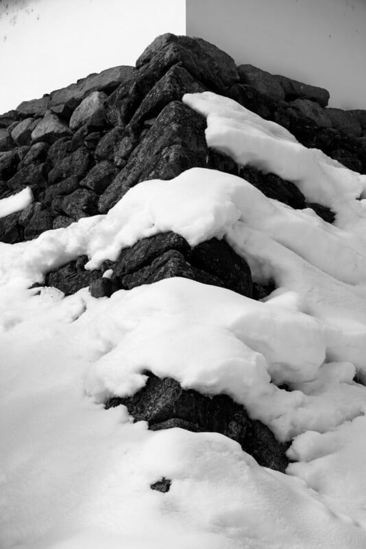 Snow-covered rocks form a rugged, ascending path leading towards a white wall backdrop. The scene is black and white, highlighting the contrast between the dark stones and the bright snow.