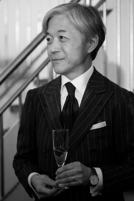 A person in a pinstripe suit holds a champagne glass, smiling slightly and looking to the side. They have short, gray hair and wear a watch. The background features a staircase with metal railings. The image is in black and white.