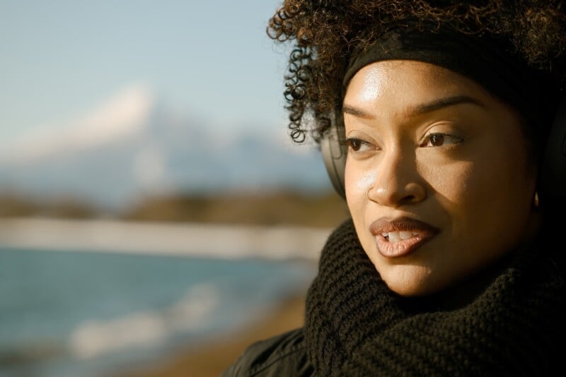 Woman with curly hair and headphones stands near a body of water, wearing a black scarf and headband. She gazes thoughtfully into the distance, with a snow-capped mountain in the blurred background.